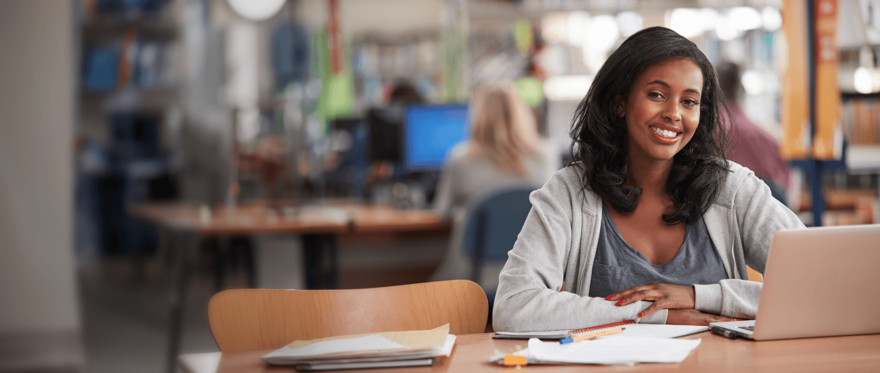 A young black woman is sitting in a library with her laptop and notebooks.