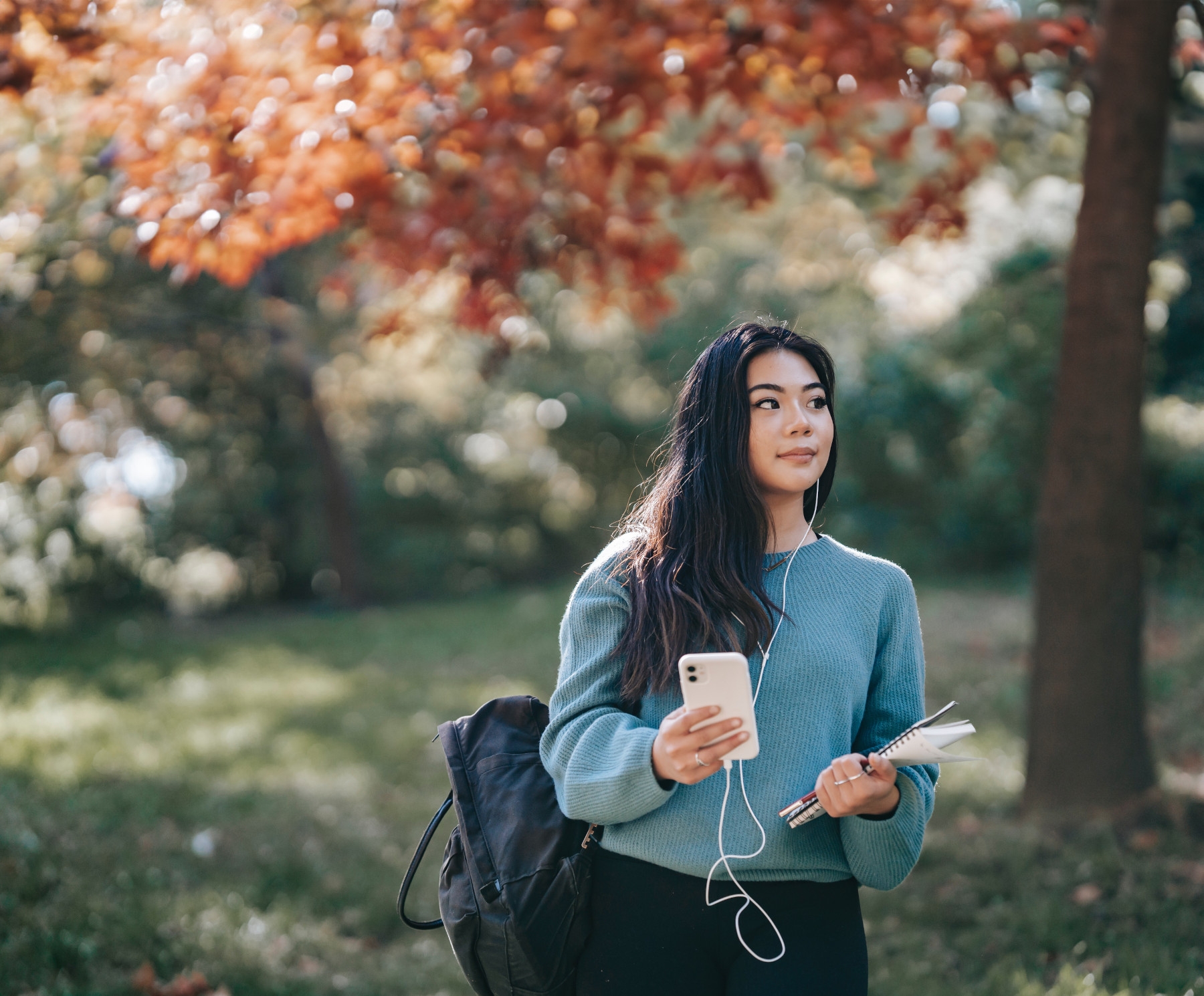 A female student is outside on her phone carrying her backpack and notebooks.