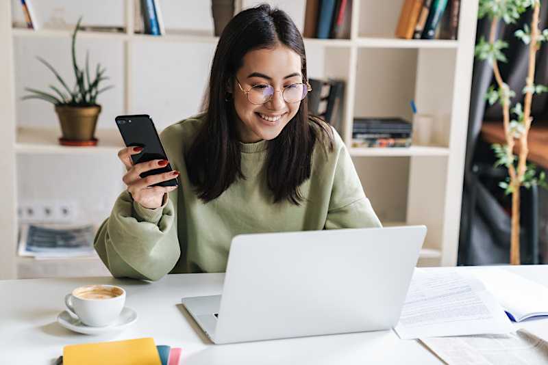 A young woman is at a desk looking at a laptop and holding her phone, smiling. 