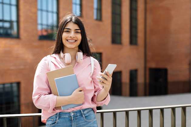 A female college student has her books and headphones on. She is smiling and looking at the camera. 