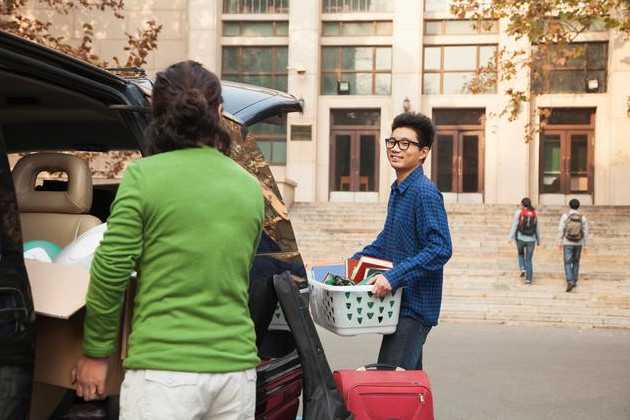 There is a woman standing at the back of an open vehicle that is packed with belongings.  A young man is carrying some belongings into a college building smiling back at his mother.