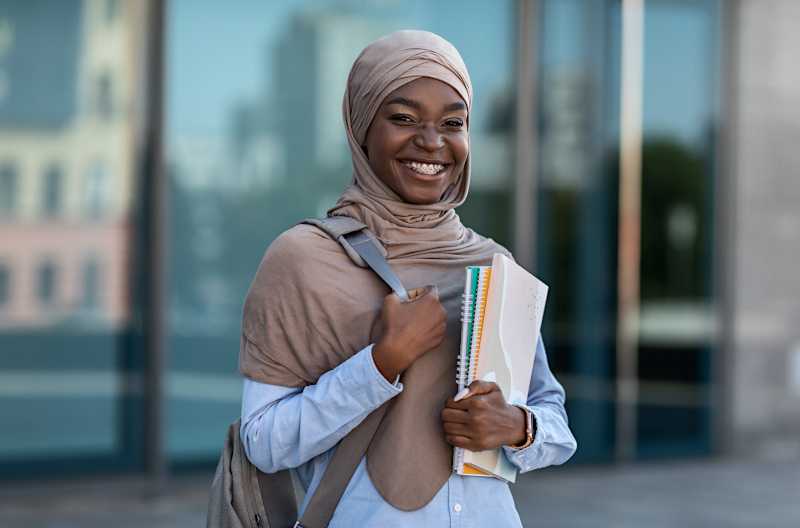 A black student wearing a hijab and carrying books smiles at the camera. 